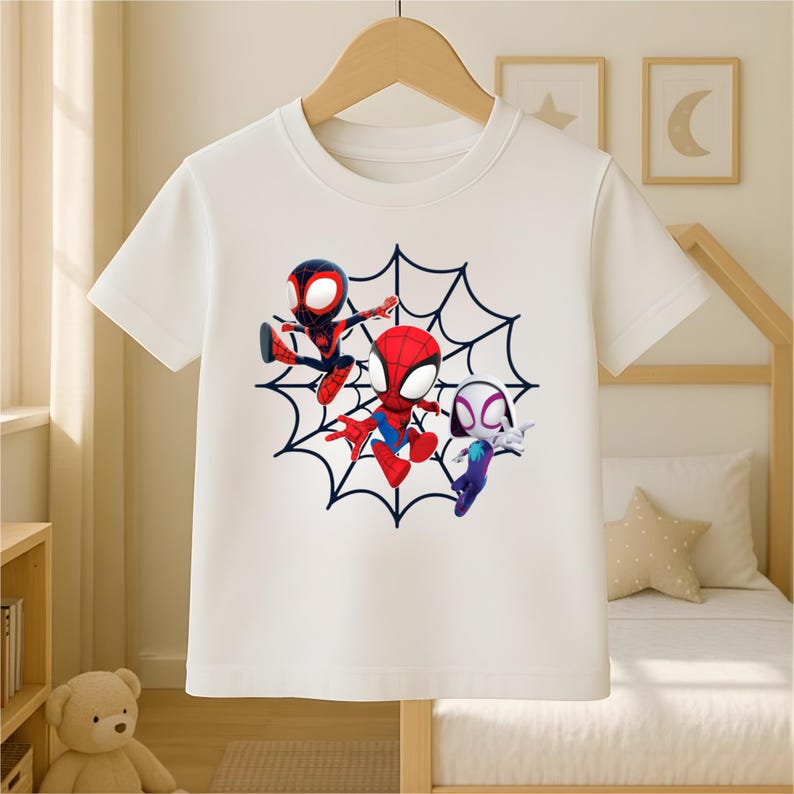 May include: White t-shirt featuring three cartoon Spider-Man characters in red, black, and blue, set against a black spiderweb design. The shirt is hanging on a wooden hanger.