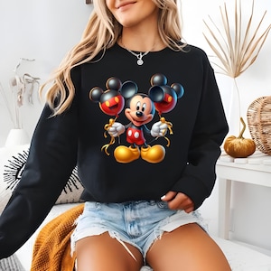 May include: Black sweatshirt featuring a vibrant Mickey Mouse graphic holding two Mickey Mouse-shaped balloons. The balloons are red and black, with gold ribbon. Mickey Mouse is wearing red shorts, white gloves, and yellow shoes.