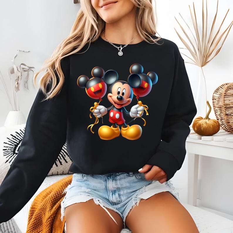 May include: Black sweatshirt featuring a vibrant Mickey Mouse graphic holding two Mickey Mouse-shaped balloons. The balloons are red and black, with gold ribbon. Mickey Mouse is wearing red shorts, white gloves, and yellow shoes.