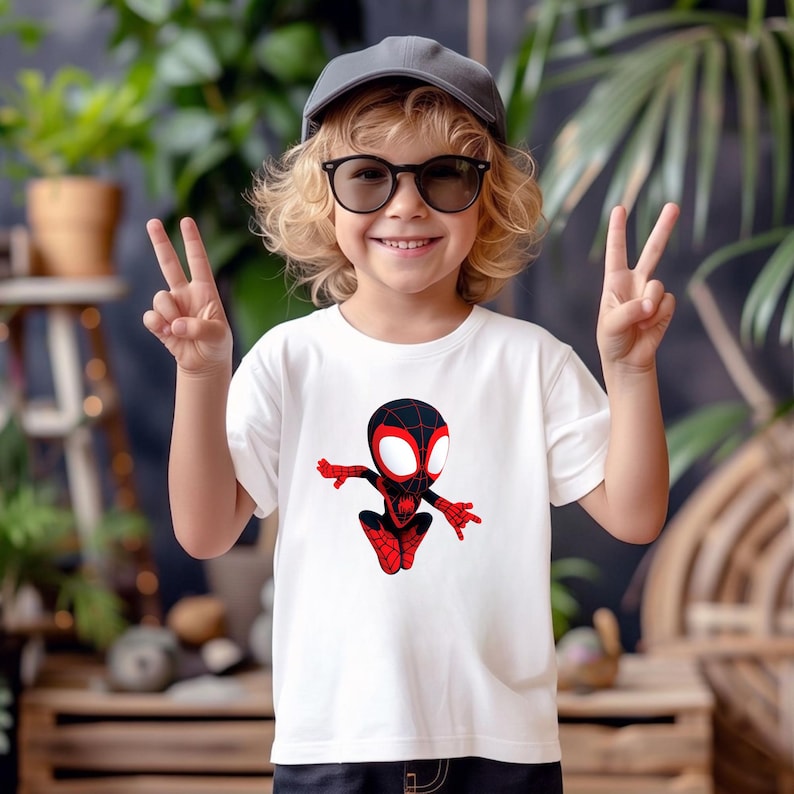 May include: White t-shirt featuring a cartoon graphic of a superhero in red and black. The superhero has large white eyes and is in a jumping pose. The shirt is worn by a child wearing a gray cap and black glasses.
