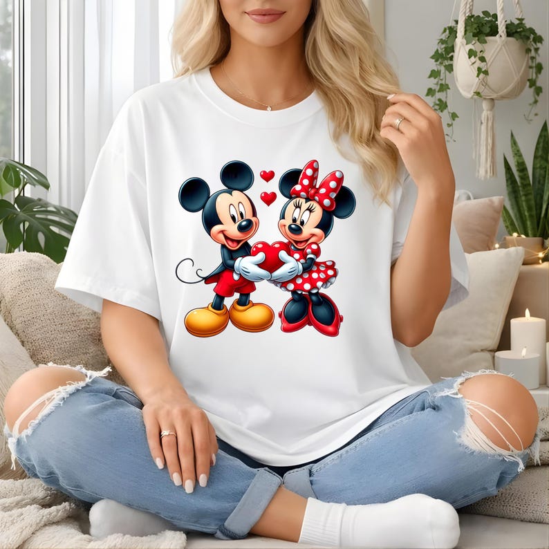 May include: White t-shirt featuring a colorful graphic of Mickey and Minnie Mouse holding a red heart. Mickey is wearing red shorts and yellow shoes, while Minnie is in a red polka-dot dress. The design includes red heart accents.