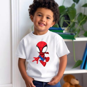 May include: A white t-shirt featuring a cartoon Spider-Man graphic. The superhero is depicted in red and blue, with large white eyes. The shirt is worn by a young child.