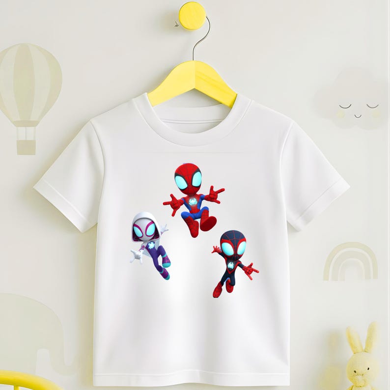May include: White t-shirt featuring three cartoon Spider-Man characters in red, blue, black, and white. The characters are in a dynamic pose, with a yellow hanger in the background. The shirt is made of a soft material.