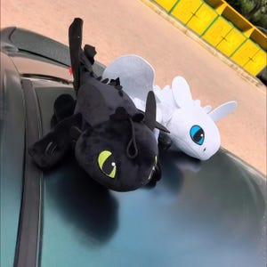 Puede incluir: Dos juguetes de peluche con forma de dragón, uno negro con ojos verde brillante y otro blanco con ojos azul claro. Ambos cuentan con cabezas grandes, orejas puntiagudas y alas pequeñas, inspirados en los personajes de la famosa franquicia.