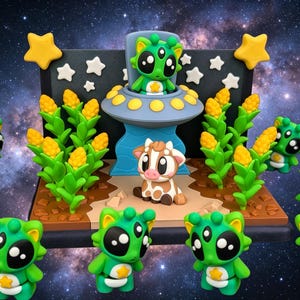 Puede incluir: Un diorama caprichoso con figuras de extraterrestres verdes, una vaca y un OVNI. La escena incluye mazorcas de maíz, estrellas amarillas y blancas sobre un fondo cósmico. Los extraterrestes tienen grandes ojos negros y detalles en forma de estrella.