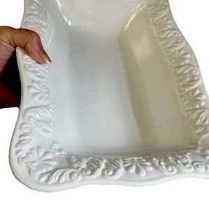 May include: A rectangular white ceramic serving tray featuring a raised, ornate relief pattern along the scalloped edges. The surface of the tray is smooth and glossy, with a deep centre basin and a decorative border of embossed floral motifs.
