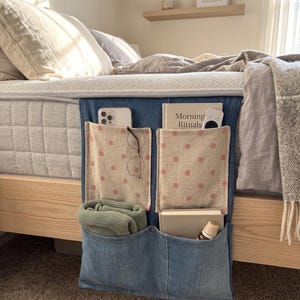 May include: A denim bed organizer with multiple pockets. The pockets are lined with a floral patterned fabric and hold items like a phone, glasses, a book titled "Morning Rituals", and a rolled-up green sweater. The organizer hangs from the bed frame.