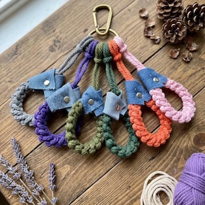 May include: A set of five wristlet keychains attached to a brass-toned metal carabiner. Each keychain features a braided cord in gray, purple, olive green, orange, or pink, accented with a blue denim fabric tab secured by a silver metal rivet.