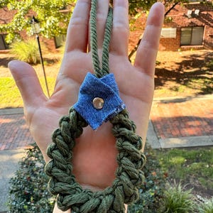 Può includere: Un portachiavi circolare in corda intrecciata verde oliva con un dettaglio in tessuto denim blu sulla parte superiore. Il pezzo di denim è piegato a forma triangolare e fissato con un bottone a pressione in metallo argentato. Un passante in corda verde abbinato si estende verso l'alto.