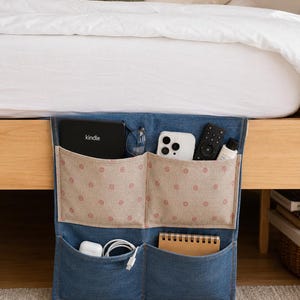 May include: A blue denim bedside storage organizer featuring two upper pockets with a beige fabric overlay decorated with small red floral patterns and two lower denim pockets. The organizer holds a Kindle, smartphone, remote, and cables.