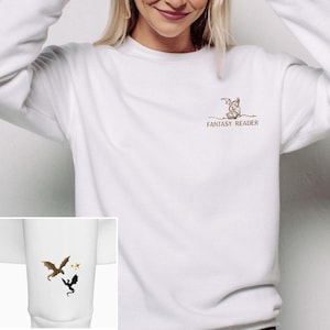 Könnte beinhalten: Weißes Sweatshirt mit einem kleinen gestickten Design eines Drachen, der ein Buch liest, mit dem Text "FANTASY READER" darunter. Ein kleineres Bild zeigt einen Drachen und Sterne. Das Sweatshirt hat einen Rundhalsausschnitt und lange Ärmel.