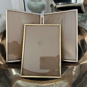 May include: A set of three gold-toned picture frames, one rectangular and two hinged, displayed in a silver-colored bowl. The frames have a simple, elegant design, perfect for displaying photos or artwork.
