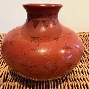 May include: A terracotta-colored ceramic vase with a rounded body and a narrow neck. The vase features hand-painted black geometric and swirling designs. The vase is displayed on a woven wicker surface.