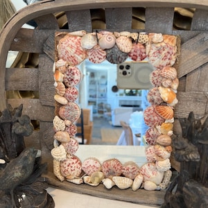 May include: A rectangular mirror with a frame decorated with various seashells. The mirror is set within a rustic, woven wooden basket. Two dark, decorative sculptures flank the mirror.
