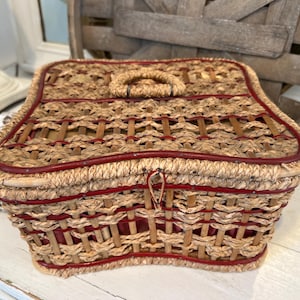 May include: A woven wicker basket with a red trim and handle. The basket has a rectangular shape with a curved top and a clasp closure. The basket is made of natural-colored wicker with red accents.