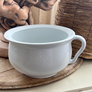 May include: A white ceramic chamber pot with a handle. The pot is round with a wide rim and sits on a wooden surface. A decorative statue and a woven basket are in the background.