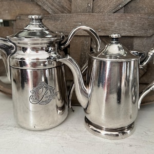 May include: Two silver-toned metal coffee pots with lids and curved handles. The larger pot features a decorative emblem with the script text Fairmont. Both vessels have a polished metallic finish, a spout, and a circular base for stability.