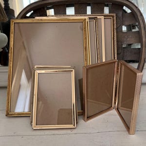 May include: A collection of gold-toned metal picture frames in various sizes, including several rectangular single frames and one hinged double frame. Each frame features a thin, textured border and contains a plain, light brown backing board.