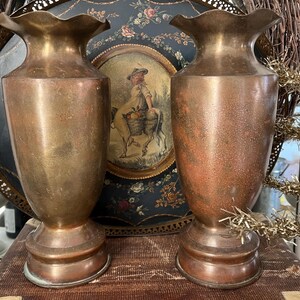 May include: Two antique brass vases with a fluted rim and a wide base. The vases have a warm, aged patina, with tones of gold and copper. They are displayed in front of a decorative tray and a painting.
