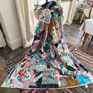 May include: A colorful patchwork quilt with a variety of fabric patterns and colors, including floral, plaid, and solid hues. The quilt is draped over a chair, showcasing its intricate design and craftsmanship. The quilt's edges are finished with a white border.
