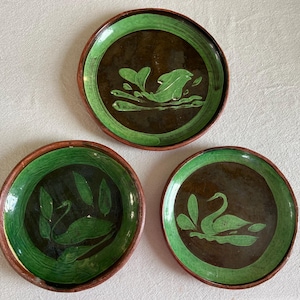 May include: Three ceramic plates with a brown center and a green border. Each plate features a green swan design. The plates have a rustic, handmade appearance, suitable for decorative or serving purposes.