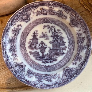 May include: A round, antique white and purple plate with a detailed Asian-inspired design. The plate features pagodas, bridges, and floral patterns. The rim has a scalloped edge.