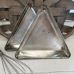 May include: Two triangular metal baking pans with a metallic silver finish and visible rust along the edges. The pans feature a flat base and raised sides, designed for shaping baked goods. A wire whisk with a wooden handle is positioned nearby.