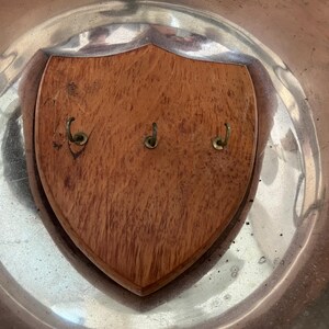 May include: A wooden key holder with a shield shape, featuring three brass hooks. The key holder is mounted on a silver-colored metal surface, creating a contrast in textures and colors. The wood grain is visible.