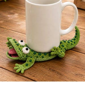 Pode incluir: Uma caneca de cerâmica branca sobre um porta-copos de crochê em forma de lagarto verde. O lagarto tem manchas verde-escuras, uma boca bem aberta com uma língua rosa e dois olhos brancos com pupilas pretas. A cauda curva-se para cima.