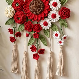 Può includere: Una decorazione da parete con fiori all'uncinetto rossi, bianchi e color crema con foglie verdi, appesa a un ramo di legno orizzontale tramite cordini color crema. La composizione presenta cinque lunghe nappe color crema pendenti.