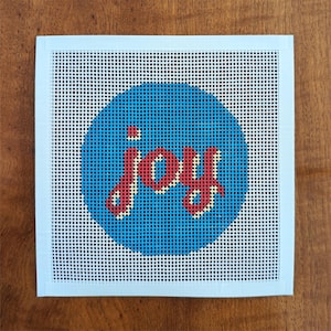 Könnte beinhalten: Ein quadratisches Stickleinen mit einem leuchtend blauen Kreis in der Mitte. Im Kreis ist das Wort joy mit rotem Garn und einer hellgelben Umrandung gestickt. Das Leinen hat einen weißen Rand und ein durchgehendes Gittermuster im Design.