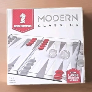 May include: A square box for a Modern Classics backgammon game set. The white packaging features a red shield logo with a white knight icon, the text Backgammon, and the words Modern Classics. It shows a clear board with red and grey checkers.