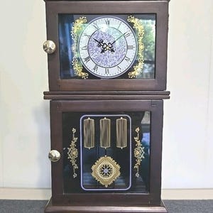 May include: A dark brown wooden mantel clock with two glass-fronted doors and a bottom drawer. The upper section features a white clock face with Roman numerals and gold floral accents. The lower section displays three gold hanging pendulums.