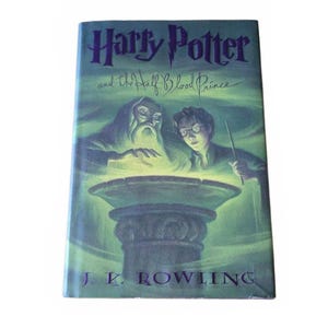 May include: A hardcover book titled Harry Potter and the Half-Blood Prince by J.K. Rowling. The cover features a green-toned illustration of an older man with a beard and a younger person with glasses, both looking into a glowing stone basin.