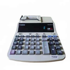 Puede incluir: Una calculadora impresora Canon P200-DH III con cuerpo de plástico gris claro, panel de pantalla negro y teclado numérico con teclas gris oscuro y gris claro. El dispositivo incluye un soporte para rollo de papel y una etiqueta de 120V.