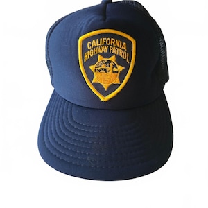 May include: A navy blue trucker cap featuring a mesh back and a curved peak. Centered on the front panel is an embroidered patch with a yellow border, yellow text reading CALIFORNIA HIGHWAY PATROL, and a central yellow star-shaped emblem.