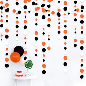 Peut inclure: Une guirlande décorative verticale composée d'une série de découpes en papier circulaires orange, noires et blanches, suspendues en plusieurs brins. Sous la guirlande, un petit gâteau aux fraises rouges est posé près d'une feuille verte et de ballons.