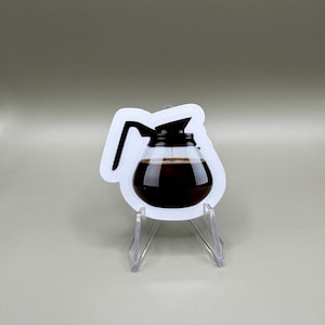May include: A sticker featuring a coffee pot filled with dark brown coffee. The coffee pot has a white outline and a black handle and lid. The sticker is displayed on a clear acrylic stand against a neutral background.