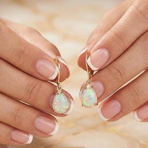 May include: A pair of gold drop earrings featuring pear-shaped opal stones. The opals show a play of colour with iridescent hues of green, blue, and yellow. The earrings are held up by a person's hands.