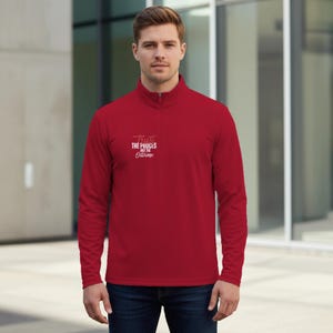 May include: A red quarter-zip pullover with the text "Trust the Process Not the Outcome" in white. The pullover has a mock neck and a zipper. The model is wearing blue jeans.