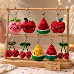 May include: A collection of nine crocheted fruit keychains hanging on a wooden stand. The keychains feature various shapes including apples, cherries, and watermelon slices, all with embroidered smiling faces and silver metal key rings.