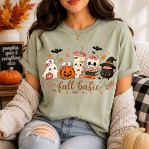May include: A sage green t-shirt featuring a graphic print of a ghost, a jack-o'-lantern, a drink cup, a skull, and a cauldron with the text fall basic. The design includes medical-themed accessories like a nurse cap, stethoscope, and books.