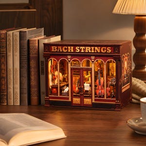 Può includere: Un modello di negozio in miniatura con la scritta "BACH STRINGS" in lettere dorate. Il negozio è di colore rosso scuro con finestre ad arco che espongono violini e altri strumenti a corda. Il modello è su una superficie di legno.