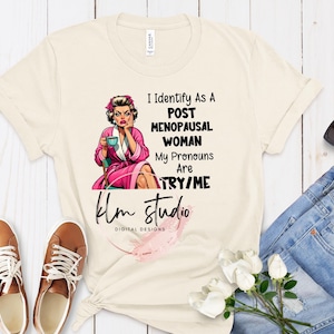 Op de afbeelding: Een beige T-shirt met een cartoonillustratie van een vrouw die een kopje vasthoudt. De tekst op het shirt luidt "I Identify As A Post Menopausal Woman My Pronouns Are TRY/ME klm studio DIGITAL DESIGNS".