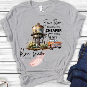 Puede incluir: Una camiseta gris con un diseño gráfico de una torre de agua oxidada, una camioneta y el texto "Back Roads Because It's Cheaper Than Therapy". El diseño está rodeado de hierba verde y flores silvestres. La camiseta también tiene un logotipo rosa acuarela que dice "klm studio digital designs".