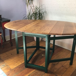 May include: A drop-leaf table featuring a natural wood grain oval top and a dark forest green painted wooden base. The table has two hinged side leaves that can be lowered or raised, supported by a sturdy frame with straight legs and crossbars.