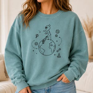 Könnte beinhalten: Ein blaugrünes Sweatshirt mit Rundhalsausschnitt und einer schwarzen Strichzeichnung eines Außerirdischen, der auf der Erde steht und einen Schraubenschlüssel hält. Das Design enthält verstreute mechanische Symbole wie Zahnräder, eine Schraube und einen Pylon.