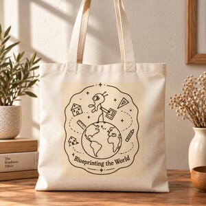 Puede incluir: Una bolsa de tela color crema con una ilustración de líneas en marrón oscuro de una figura de palo sobre un globo terráqueo. El diseño incluye iconos de casas, una regla, un lápiz y una escuadra, con el texto "Blueprinting the World" debajo.