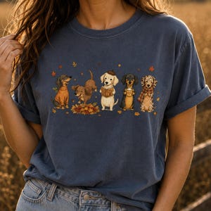 May include: A slate blue short-sleeve t-shirt featuring a graphic print of five dachshund dogs in autumn-themed attire. The dogs are depicted playing in fallen leaves, reading a book, holding a mug, and wearing sweaters and scarves.