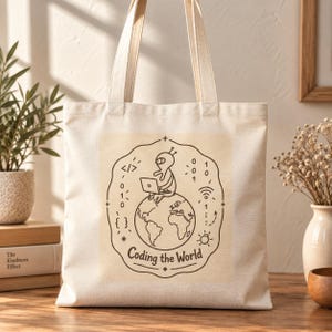 May include: A beige canvas tote bag featuring a dark brown line art graphic of an alien sitting on a globe while using a laptop. The design includes binary code, a Wi-Fi symbol, and the text Coding the World enclosed in a scalloped border.